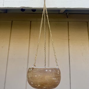 May include: A hanging planter with a speckled, light brown ceramic pot suspended by a gold-colored chain. The pot has a rounded shape and a white interior. The chain is attached to a metal ring at the top.