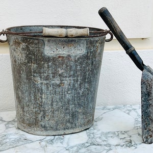 May include: A vintage galvanised metal bucket with a wooden handle and a matching metal scoop. The bucket and scoop both show signs of wear and tear, giving them a rustic look.
