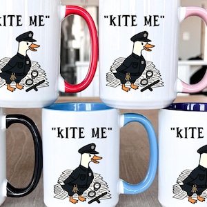 May include: White ceramic mugs with colorful handles and rims. Each mug features the text "KITE ME" and a cartoon duck in a police uniform. The mugs have black, red, pink, blue, and navy blue accents.