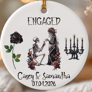 May include: A round white ceramic ornament hanging from a gold string. It features the text ENGAGED, Casey & Samantha, and 07.04.2026. The graphic shows a skeleton proposing to a skeleton bride, a black rose, and a black candelabra.