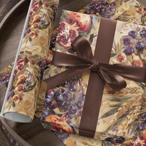 Puede incluir: Caja de regalo envuelta en papel decorativo con un diseño de frutas y trigo, atada con una cinta marrón. También se ve un rollo de papel de regalo a juego y una hoja del mismo diseño. El diseño presenta granadas, uvas e higos.