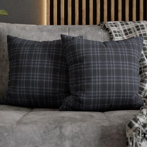 May include: Two square cushions with a dark grey plaid pattern, placed on a grey sofa. A matching plaid throw blanket is draped over the sofa. The cushions and blanket add a touch of pattern and texture to the neutral-toned living space.