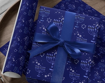 Puede incluir: Una caja de regalo rectangular envuelta en papel azul oscuro con un patrón repetitivo blanco de menorás, estrellas y copos de nieve. La caja está atada con un lazo de cinta de satén azul oscuro. Un rollo de papel a juego se encuentra al lado.