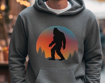 Peut inclure: Un sweat à capuche en gris chiné anthracite avec un motif représentant un Bigfoot marchant devant un coucher de soleil. Le motif est dans un cercle avec des arbres de chaque côté du Bigfoot.