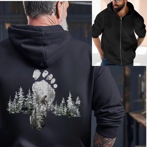 May include: Black zip-up hoodie with a Bigfoot footprint design on the back. The footprint graphic features a forest scene with a silhouette of Bigfoot. The hoodie has a hood and a front zipper. The word "BLACK" is printed on the bottom right.