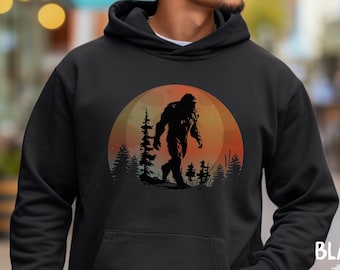 Peut inclure: Sweat à capuche noir avec un motif de Bigfoot devant un coucher de soleil et des arbres. Le mot "BLACK" est affiché en bas à droite. Le sweat-shirt a une poche avant et une capuche à cordon.
