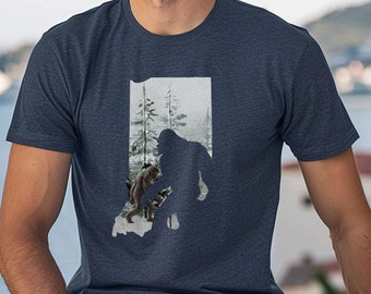 Può includere: Una t-shirt blu navy heather con una stampa grafica di Bigfoot e un Bigfoot più piccolo che cammina in una foresta. La grafica ha la forma dello stato dell'Indiana.