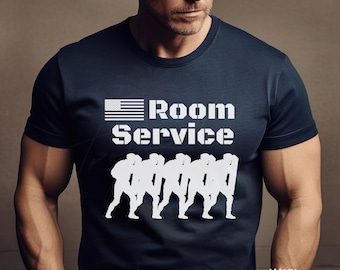 May include: A navy blue t-shirt featuring a white American flag, the words "Room Service" and a silhouette of a group of people in military uniform. The shirt is made from a soft, comfortable material and is ideal for everyday wear.