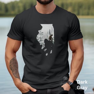 May include: A black t-shirt with a white outline of the state of Illinois. Inside the outline are silhouettes of two Bigfoot figures walking through a snowy forest.