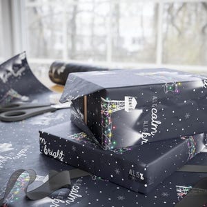 Pode incluir: Dois rolos de papel de embrulho preto e branco com um design de farol e o texto "ALL IS calm. ALL IS bright." Os rolos est&atilde;o empilhados um em cima do outro.