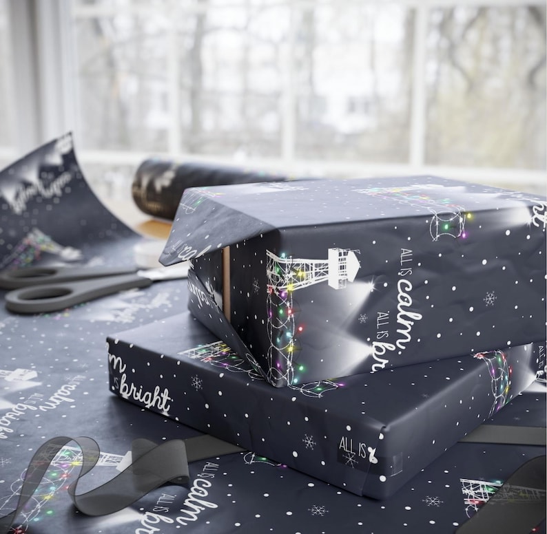 Pode incluir: Dois rolos de papel de embrulho preto e branco com um design de farol e o texto "ALL IS calm. ALL IS bright." Os rolos est&atilde;o empilhados um em cima do outro.