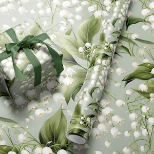 May include: A green and white gift wrap with a floral pattern of lily of the valley flowers. The gift wrap is wrapped around a gift box with a green ribbon.