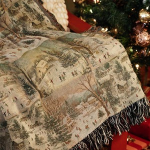 May include: A woven tapestry throw blanket with a winter scene design. The blanket features a detailed illustration of a snowy village with people, trees, and houses. The blanket has a fringed edge and is draped over a red surface.