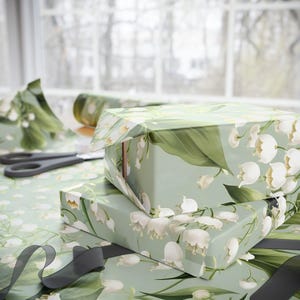 May include: Two gift boxes wrapped in green and white floral wrapping paper with a lily of the valley design. The boxes are stacked on top of each other and are tied with black ribbon.