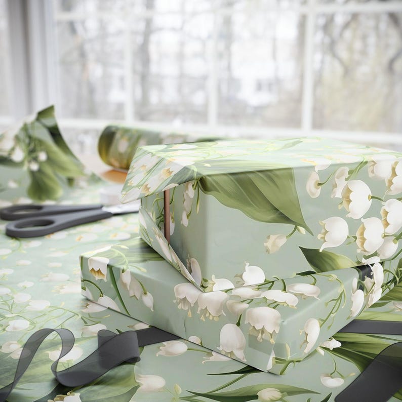 May include: Two gift boxes wrapped in green and white floral wrapping paper with a lily of the valley design. The boxes are stacked on top of each other and are tied with black ribbon.