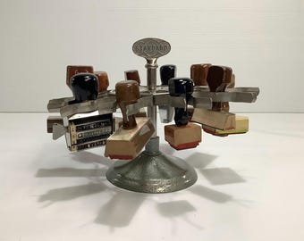 May include: A vintage metal stamp carousel with multiple stamps. The stamps have wooden handles and red ink pads. The carousel has a silver base and a "STANDARD" label.