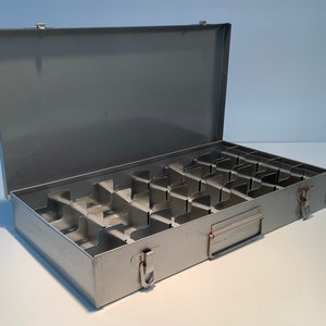 Peut inclure: Une boîte de rangement en métal argenté avec un couvercle à charnières et une poignée. La boîte comporte plusieurs compartiments à l'intérieur.