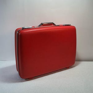 May include: A rectangular red hard-shell suitcase with a textured surface. The suitcase features a silver-toned metal handle at the top, silver-toned metal latches, and a silver-toned trim running along the perimeter of the closure.