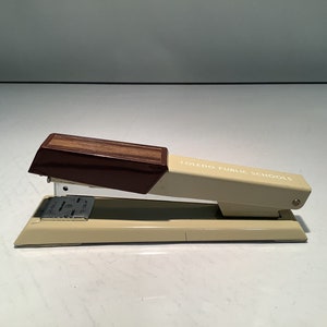 May include: A vintage beige stapler with a dark brown wooden top. The stapler has the text "TOLEDO PUBLIC SCHOOLS" printed on the side. The stapler is made of metal and plastic.