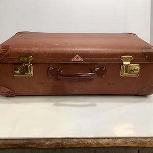 May include: A vintage brown leather suitcase with brass hardware and a leather handle. The suitcase has a small pink triangle sticker on the top.