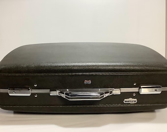 May include: A vintage dark green suitcase with silver-toned hardware. The suitcase has a rounded top, a handle, and a small emblem. The brand name "Titan" is visible on the side. A classic travel accessory.