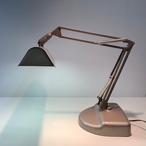 Könnte beinhalten: Eine Vintage-Schreibtischlampe mit grauem Finish. Die Lampe verfügt über einen verstellbaren Arm mit Federmechanismus und einen rechteckigen Schirm. Der Sockel hat eine abgerundete Form. Die Lampe ist für fokussiertes Licht konzipiert.