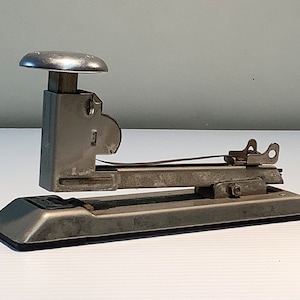 May include: A silver metal stapler with a large, round, silver top. The stapler has a long, rectangular base and a lever on the side.