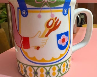 Könnte beinhalten: Weiße Keramik-Tasse mit farbenfrohem Schürzen-Design. Die Schürze zeigt eine Hand, die einen Löffel und eine Gabel hält, mit blauen, gelben und rosa Akzenten. Die Tasse hat einen weißen Henkel und einen dekorativen grün-gelben Rand.