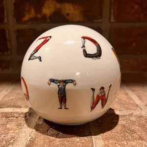 May include: White ceramic sphere with colourful illustrations of people forming letters of the alphabet. The sphere is decorated with red, blue, yellow, and green colours.