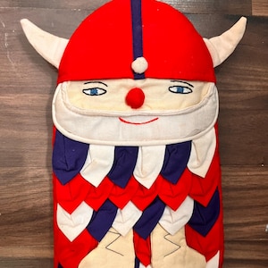 Puede incluir: Un guante de cocina de fantasía con forma de vikingo. Presenta un casco rojo con una raya azul, cuernos blancos y una cara sonriente con ojos azules y nariz roja. El cuerpo es rojo con detalles blancos, azules y crema.