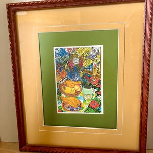 Op de afbeelding: Ingelijst kunstwerk met een kleurrijk stilleven. Het kunstwerk, geplaatst in een groene passe-partout en een goudkleurige rand, toont een vaas met bloemen, fruit en borden met patronen. De lijst is donkerbruin met een gedetailleerde rand.