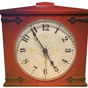 May include: A red, rectangular clock with a white face and black numerals. The clock face features a compass rose design and the words "Westclox". Black decorative hardware accents the corners. The clock hands point to 5:55.