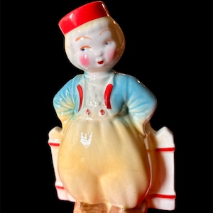 Puede incluir: Figura de cerámica vintage de un joven con vestimenta tradicional holandesa. La figura presenta un sombrero rojo, una chaqueta azul, pantalones amarillos y zuecos. La figura está agarrando una valla blanca con detalles rojos.