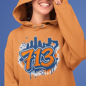 May include: Mustard-colored hoodie with a graphic featuring the number "713" in orange and blue, with a cityscape silhouette. The design includes star accents and a spray paint effect. The hoodie has a drawstring hood.