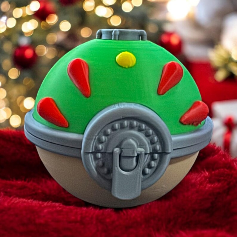 K&ouml;nnte beinhalten: Ein gr&uuml;ner und grauer Pok&eacute;ball mit roten Akzenten und einem gelben Knopf. Der Pok&eacute;ball ist geschlossen und hat einen Verschluss an der Seite.