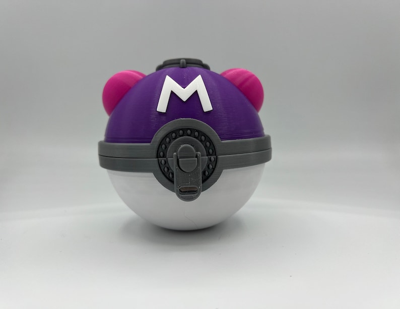 K&ouml;nnte beinhalten: Ein lila-wei&szlig;er Pok&eacute;ball mit einem rosa Knopf auf jeder Seite und einem wei&szlig;en "M" auf der Vorderseite.