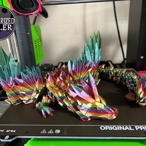 May include: A 3D-printed dragon figurine in iridescent rainbow colors. The dragon is made of geometric crystal-like shapes. The wings and body are detailed with sharp edges. The figurine is on a black surface.