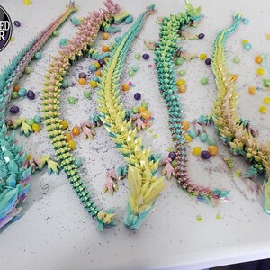 May include: Four articulated dragon figurines in pastel colors: blue, yellow, pink, and green. Each dragon is decorated with crystal-like spikes and is surrounded by colorful candies. The image includes an "AUTHORIZED SELLER" logo.