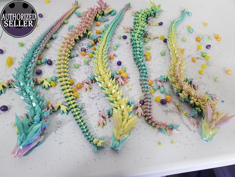 May include: Four articulated dragon figurines in pastel colors: blue, yellow, pink, and green. Each dragon is decorated with crystal-like spikes and is surrounded by colorful candies. The image includes an "AUTHORIZED SELLER" logo.