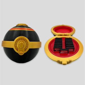 Puede incluir: Un contenedor decorativo negro y dorado, que se asemeja a una bola estilizada, con un acento rojo. El contenedor está abierto, revelando varios cartuchos de juego negros. El contenedor está diseñado para guardar cartuchos de juegos de Nintendo Switch.