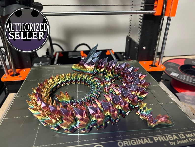 May include: A 3D-printed, articulated dragon figurine with a rainbow iridescent finish. The dragon is posed on a printer bed. The dragon's body is made of many crystal-like segments. The image includes a printer and a spool of filament.