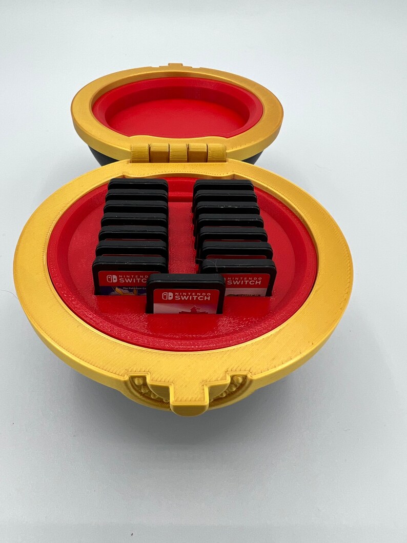 K&ouml;nnte beinhalten: Ein gold- und rotes Nintendo Switch-Spielkartuschen-Aufbewahrungsetui mit einem Scharnierdeckel. Das Etui ist kugelf&ouml;rmig und hat ein dekoratives Design. Das Etui ist ge&ouml;ffnet und zeigt 15 Spielkartuschen im Inneren.