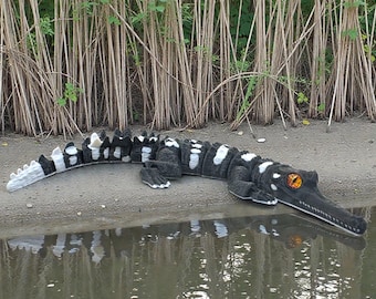 May include: A black and white alligator sculpture with orange eyes rests on a sandy bank near water. The alligator has a long snout and a textured body with white spots. Tall, dried plants form a backdrop.