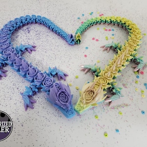 May include: Two articulated dragon sculptures, forming a heart shape. One dragon transitions from blue to purple, the other from yellow to green. Each dragon is adorned with rose-shaped details and sharp spines. The background is white with colorful confetti.