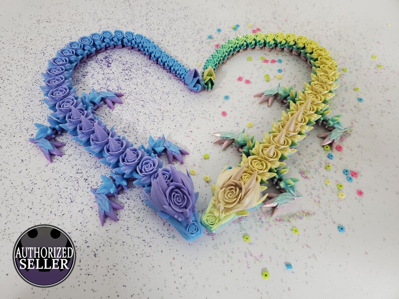 May include: Two articulated dragon sculptures, forming a heart shape. One dragon transitions from blue to purple, the other from yellow to green. Each dragon is adorned with rose-shaped details and sharp spines. The background is white with colorful confetti.