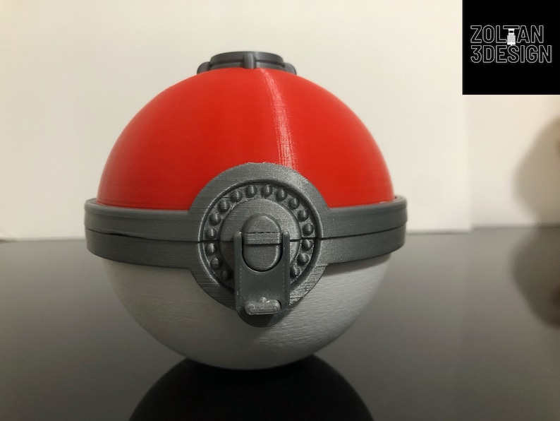 K&ouml;nnte beinhalten: Ein 3D-gedruckter Pok&eacute;ball mit roter Oberseite, wei&szlig;er Unterseite und einem grauen Verschluss. Der Pok&eacute;ball hat einen grauen Ring um die Mitte mit einer Reihe kleiner grauer Kreise.