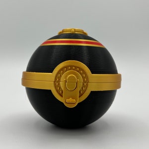 K&ouml;nnte beinhalten: Eine schwarze Kugel mit einem goldenen Band und einem roten Streifen. Die Kugel hat einen goldenen Verschluss und ein dekoratives Muster.
