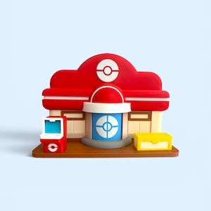 Op de afbeelding: Een miniatuur rood en wit gebouw met een blauw-witte deur, een rode arcade machine en een gele doos. Het gebouw heeft een bruine basis en een rood dak met een wit cirkelontwerp. De arcade machine is rood met een blauw scherm.