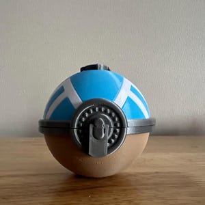 K&ouml;nnte beinhalten: Ein blauer und wei&szlig;er Pok&eacute;ball mit einem grauen Verschluss. Der Pok&eacute;ball ist aus Plastik und hat eine braune Basis.