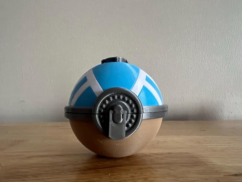 K&ouml;nnte beinhalten: Ein blauer und wei&szlig;er Pok&eacute;ball mit einem grauen Verschluss. Der Pok&eacute;ball ist aus Plastik und hat eine braune Basis.